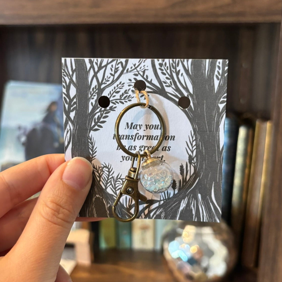 Hand holding a keychain card with a tree design in front of a bookshelf.